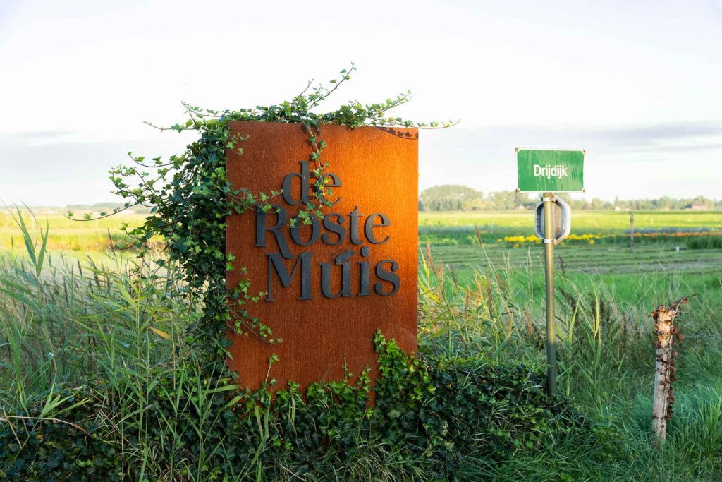 zomers sfeerbeeld van het zonovergoten terras van restaurant Roste Muis in Waterland-Oudemand bekend om z’n palinggerechten. Het restaurant bevindt zich aan Oudemanskreek vlakbij het bekende kunstwerk van Chris Ferket met de naam “Als de vos de passie preekt boer let op uw ganzen.” Het kunstwerk  staat symbool voor het Meetjeslandse krekengebied.
