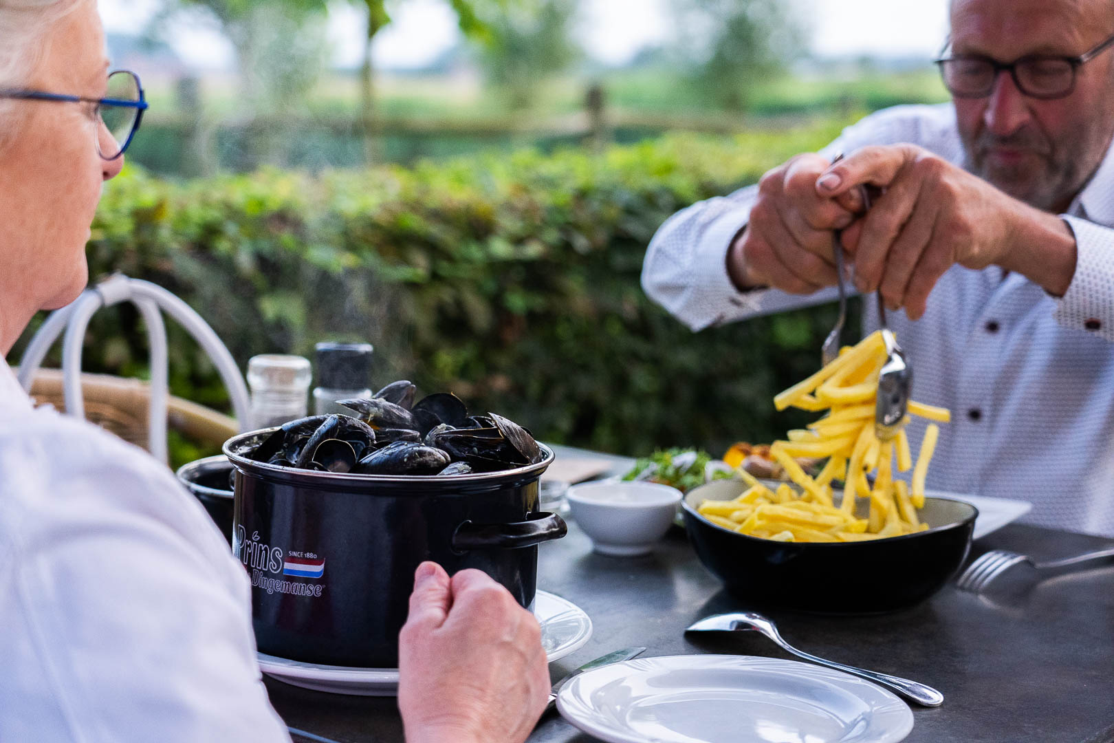 zomers sfeerbeeld van het zonovergoten terras van restaurant Roste Muis in Waterland-Oudemand bekend om z’n palinggerechten. Het restaurant bevindt zich aan Oudemanskreek vlakbij het bekende kunstwerk van Chris Ferket met de naam “Als de vos de passie preekt boer let op uw ganzen.” Het kunstwerk  staat symbool voor het Meetjeslandse krekengebied.